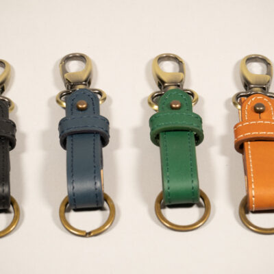 Présentation des porte-clefs ICONIK, alignés sur un fond photo gris. Les portes-clefs reprennent les colroris de la collection ICONIK, de gauche à droite: Noir, Bleu pétrole, Vert sapin et camel avec une bouclerie de couleur laiton vieilli.