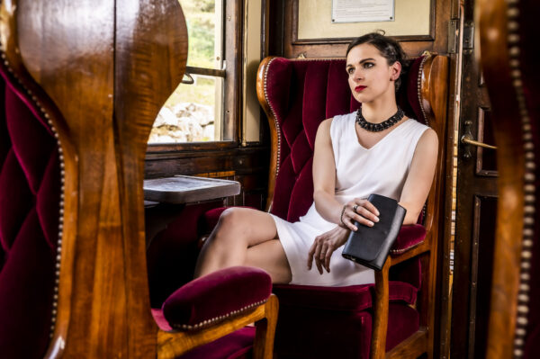 Sac 2 en 1 noir porté à la main du mannequin, assise dans un wagon de train ancien,sur un fauteuil d velours rouge matelassé.