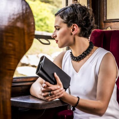 Sac 2 en 1 noir de la collection Intemporels, porté à la main du mannequin, assise dans un wagon de train ancien,sur un fauteuil d velours rouge matelassé.